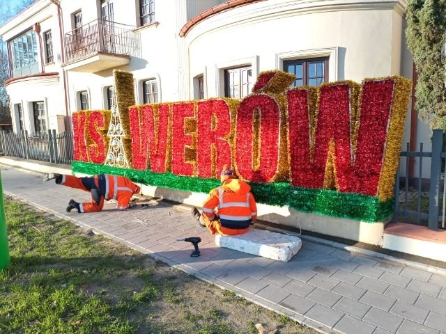 Czerwono- złoty napis KSAWERÓW z choinką zamiast litery A. Obok pracownicy, którzy montują napis na ogrodzeniu Domu Kultury