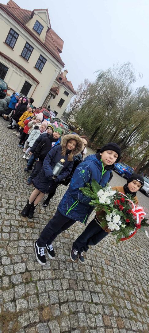 Grupa dzieci jedno z nich trzyma znicz by zapalić i złożyć pod pomnikiem.