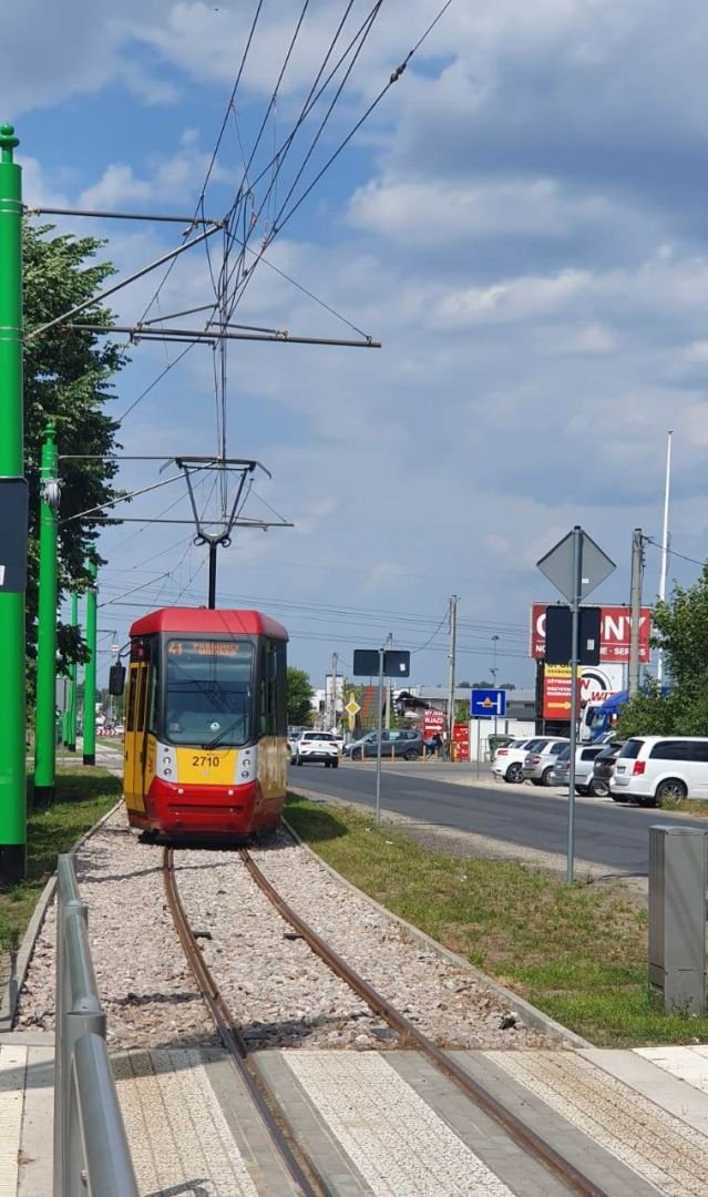 Czerwono-żółty tramwaj na torowisku. W tle samochody zaparkowane na poboczu jezdni