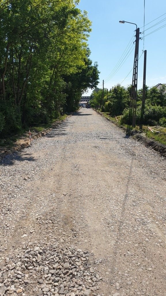 Zdjęcie przedstawia ulicę na której wysypano kruszywo pod nakładkę asfaltową. Z lewej strony widoczne przydrożne drzewa, z prawej strony latarnia
