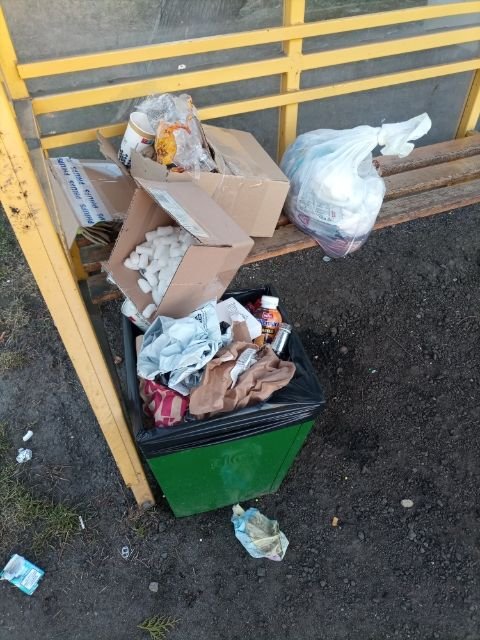 widoczny żółty przystanek autobusowy obok którego stoi zielony kosz na śmieci. Z kosza na śmieci wysypują się odpady. Na ławce przystanku leżą puste kartony po paczkach oraz pełny worek śmieci.
