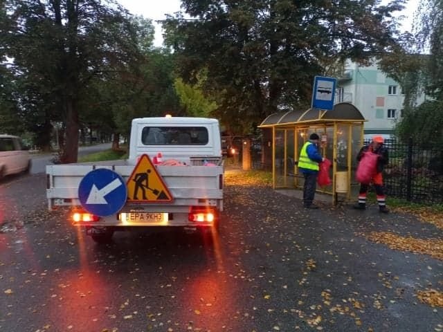 Samochód stojący koło przystanku autobusowego dwóch pracowników opróżnia kosz na śmieci. 