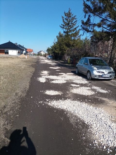 Droga gruntowa wysypana białym tłuczniem. W tle widoczne domy i drzewa