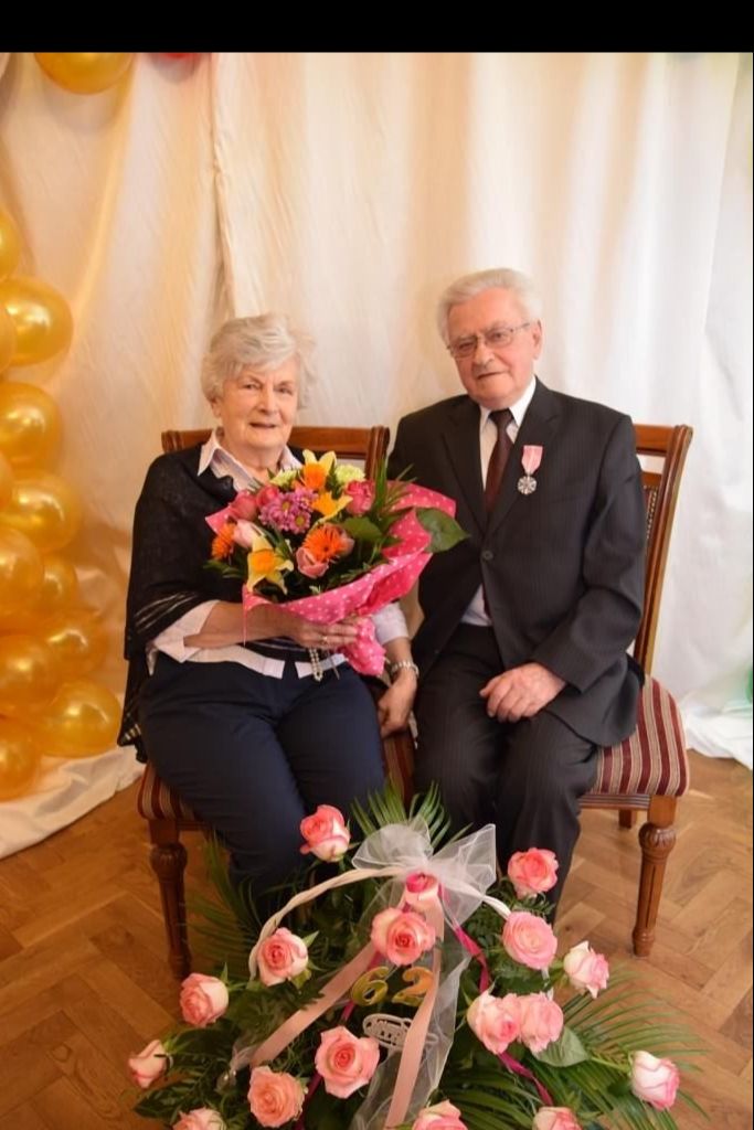 Para seniorów siedzi na krzesłach - za nimi dekoracja z balonów. Przed parą stoi kosz z kwiatami