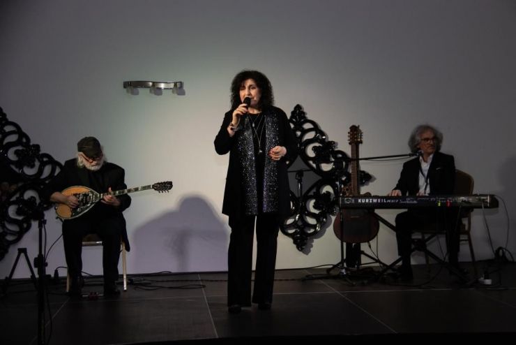 wokalistka ubrana na czarno stoi na scenie z mikrofonem. W tle muzycy z instrumentami
