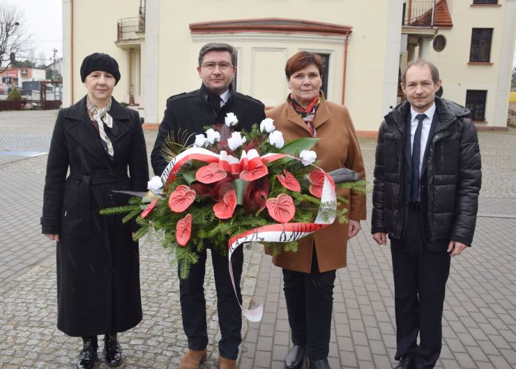 Cztery osoby stoją przed obeliskiem. Mężczyzna trzyma w dłoniach wieniec z biało czerwonymi kwiatami.