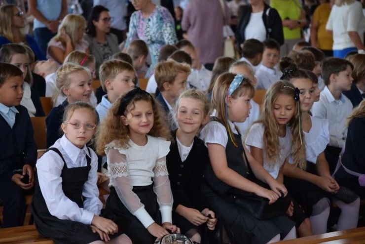 Na sali gimnastycznej siedzi grupa ubranych na galowo dzieci podczas rozpoczęcia roku szkolnego