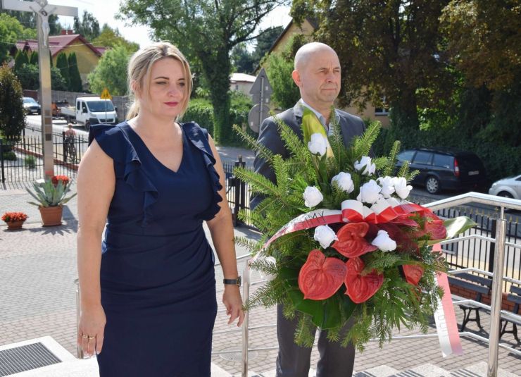 Kobieta w granatowej sukience i mężczyzna stoją na schodach przed kościołem. Mężczyzna trzyma wiązankę z biało-czerwoną szarfą