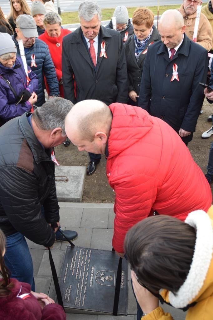 Dwóch mężczyzn umieszcza tablicę pamiątkową w miejscu płyty chodnikowej