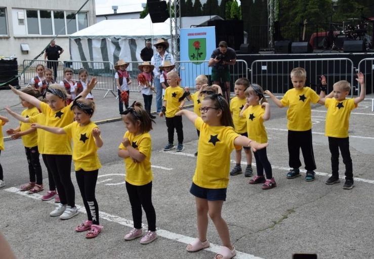 Pod sceną stoi grupa dzieci ubranych w żółte koszulki z naszytymi gwiazdkami. Dzieci ubrane są w czarne spodnie. Wszyscy mają uniesione w górę ręce.