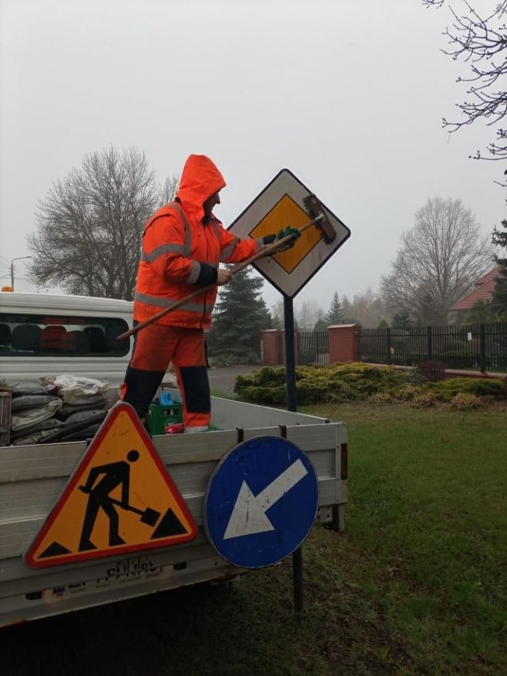Mężczyzna w pomarańczowym stroju roboczym stoi na przyczepie auta. W ręku trzyma szczotkę, którą czyści znak pionowy.