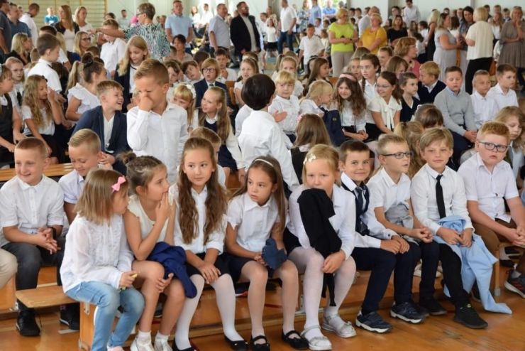 Na sali gimnastycznej siedzi grupa ubranych na galowo dzieci podczas rozpoczęcia roku szkolnego