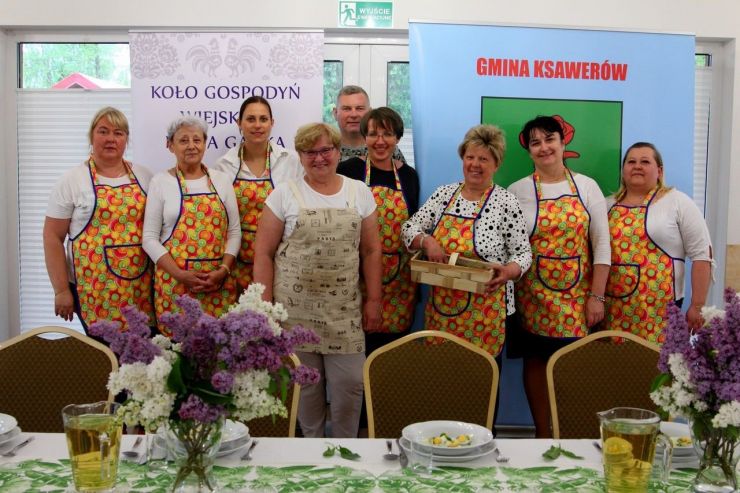 Na zdjęciu przedstawiciele KGW Nowa Gadka. 8 kobiet i 1 mężczyzna w kolorowych fartuszkach stoją na tle baneru z herbem gminy. Przed nimi udekorowany stół