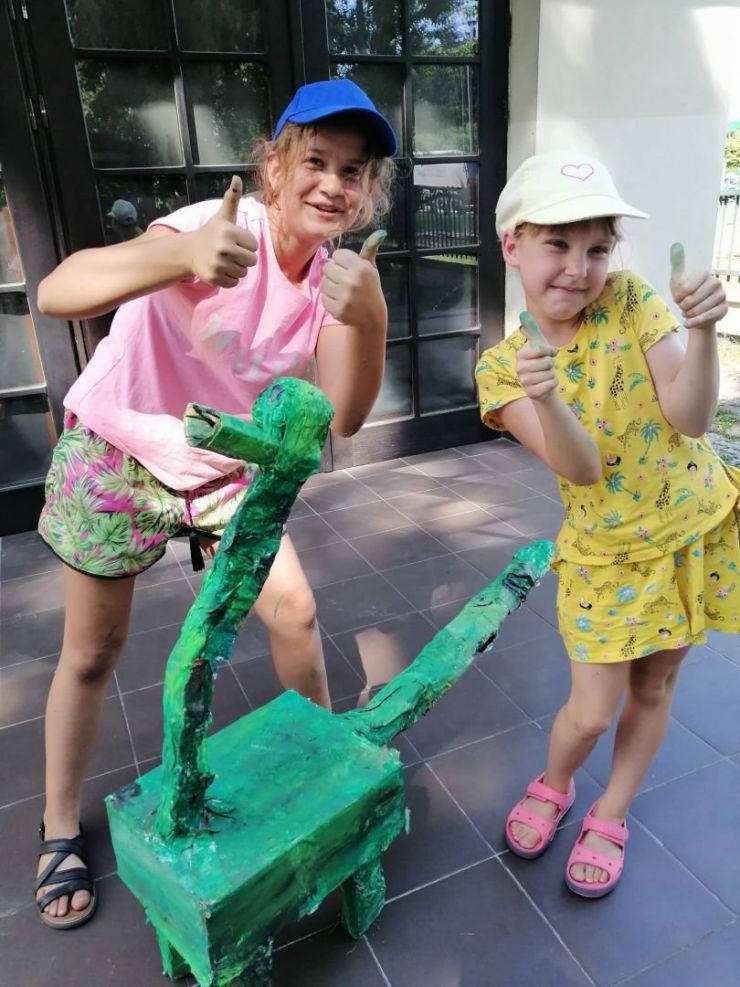 Dwie dziewczynki uśmiechają się do zdjęcia, przed mini stoi wykonany przez nie dinozaur z kartonu pomalowany na zielono.