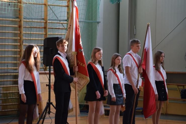 Na sali gimnastycznie stoi 2 uczniów trzymających sztandary. Jeden szkoły, drugo flagę Polski. Obok uczniów - w poczcie sztandarowym stoją cztery uczennice. Wszyscy są przepasani biało czerwoną szarfą.