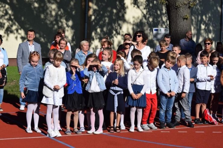 Grupa dzieci ubranych na galowo stoi na boisku podczas rozpoczęcia roku szkolnego