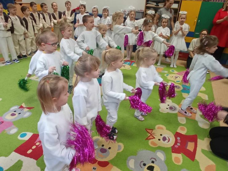 Dzieci ubrane w białe stroje trzymają w rękach kolorowe pompony. W tle inna grupa ubrana w beżowe stroje z kamizelkami