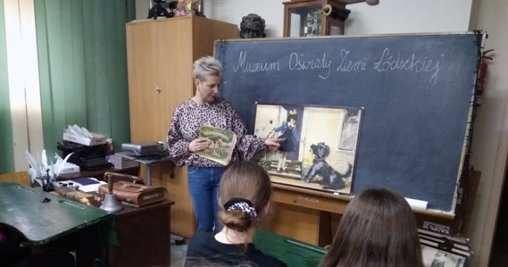 Kobieta stoi przy czarnej tablicy szkolnej na której zamocowany jest rysunek. Obok na biuru stoją różne rekwizyty