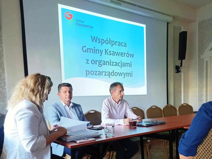 Przy stole siedzi dwóch mężczyzn i kobieta. W tle projektor z napisem "Współpraca Gminy Ksawerów z organizacjami pozarządowymi"