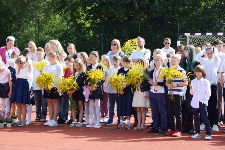 Grupa dzieci z rodzicami stoją na boisku niektóre trzymają w dłoniach bukiety kwiatów.