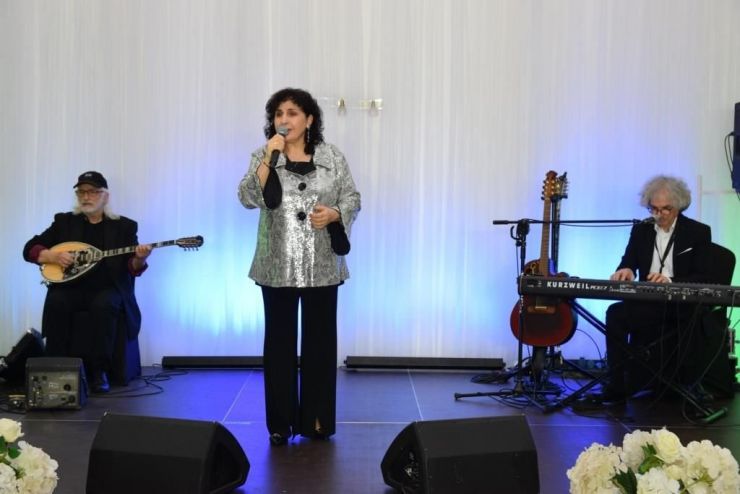 Kobieta w srebrnej bluzce stoi na scenie z mikrofonem w dłoni. Z tyłu widoczne instrumenty muzyczne