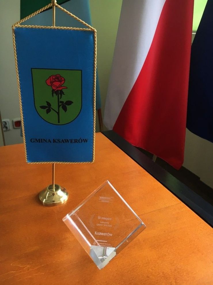szklana statuetka z napisem III miejsce kategoria gminy wiejskie Ksawerów stojąca na stole po lewej stronie flagietka gminna