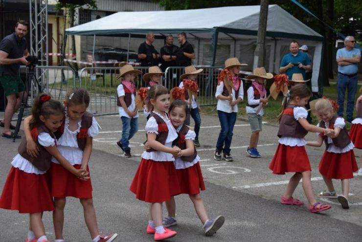 Grupa dzieci tańczy w parach. Dziewczynki ubrane są w czerwone spódniczki i brązowe marynarki z frędzlami. Chłopcy mają na głowach kapelusze i trzymają w ręku głowy koni