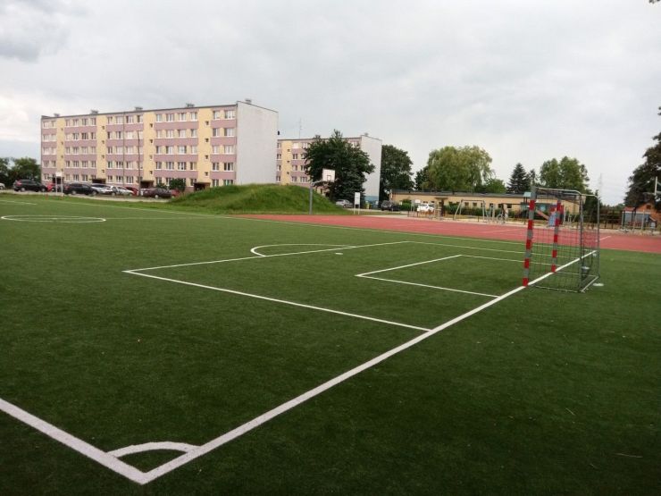 boisko z liniami do gry w piłkę nożną jedna bramka w tle dwa bloki mieszkalne i płaski budynek w którym mieści się przedszkole gminne