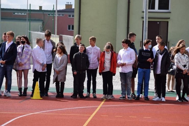 Uczniowie stoją na boisku podczas rozpoczęcia roku szkolnego. W tle widać siatkę piłkochwytu i budynek szkoły
