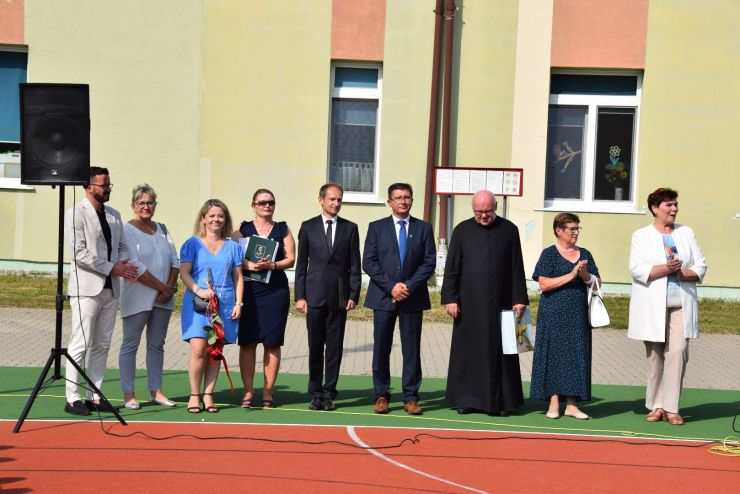 Władze gminy z zaproszonymi gośćmi stoją podczas rozpoczęcia roku szkolnego.