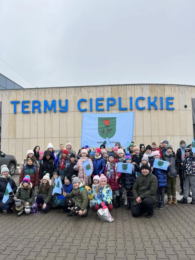 Na zdjęciu grupa dzieci w zimowych ubraniach stoi obok budynku na którym jest napis Termy Cieplice. Dzieci trzymają w rękach flagi Gminy Ksawerów