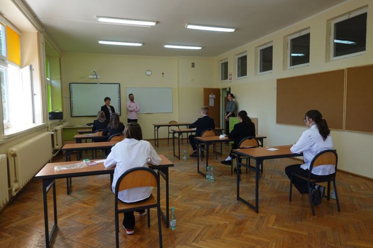 Trójka nauczycieli stoi z przodu sali. Uczniowie siedzą w ławkach podczas egzaminu.