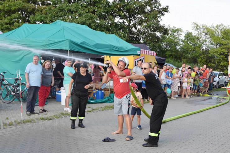 Mężczyzna w pomarańczowej koszulce trzyma w ręku węża strażackiego. Obok stoi strażak i wskazuje gdzie należy celować. W tle publiczność