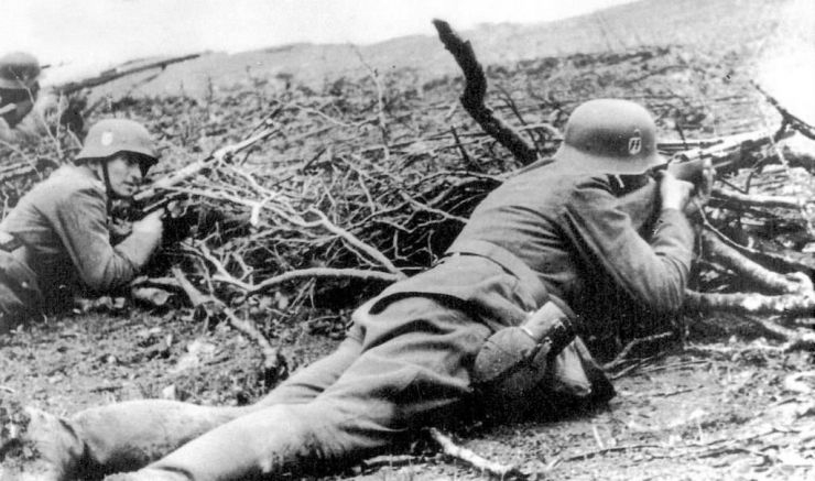 Czarno-białe zdjęcie żołnierzy z bronią. Leżą obok zasieków z gałęzi 