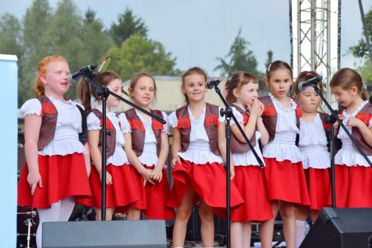 Grupa dziewcząt stoi na scenie przy mikrofonach. Dziewczynki ubrane są w czerwone sukienki, białe koszulki i brązowe marynarki z frędzlami