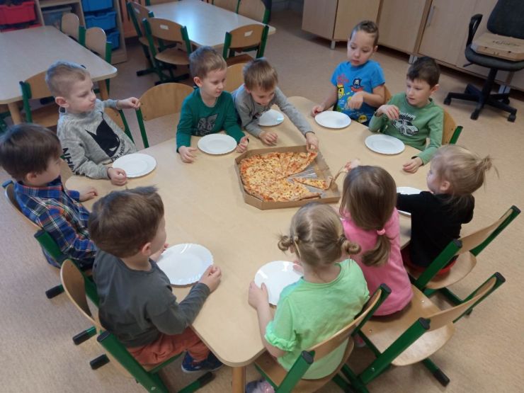 Grupa dzieci siedzi przy stoliku, na którym znajduje się pizza, którą częstują się dzieci