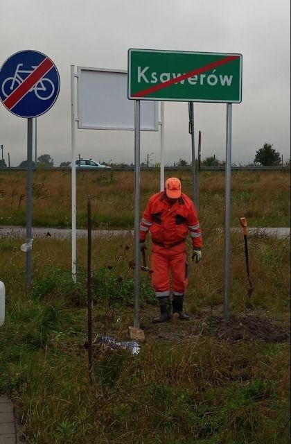 Pracownik ubrany w pomarańczowy strój służbowy stoi przy nowym oznakowaniu pionowym - czerwone przekreślenie białego napisu Ksawerów na zielonym tle.