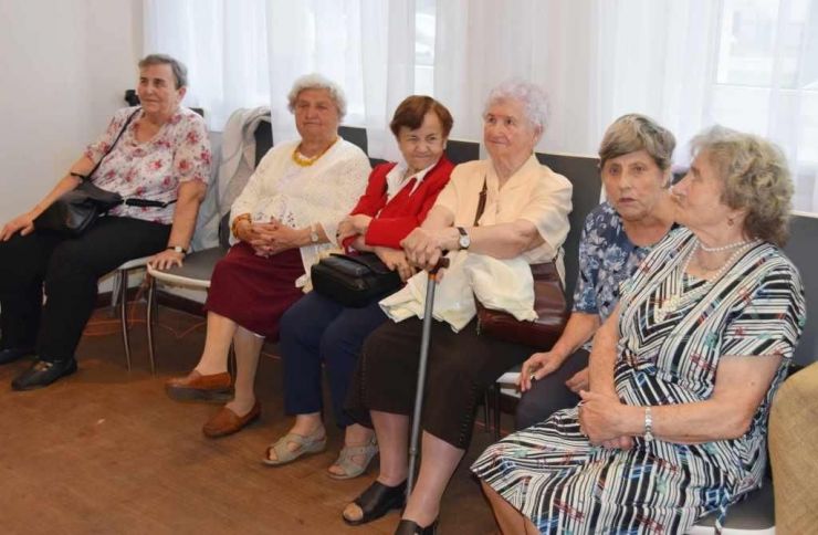 kobiety w starszym wieku, ubrane w eleganckie kreacje siedzą na krzesłach ustawionych przy oknie