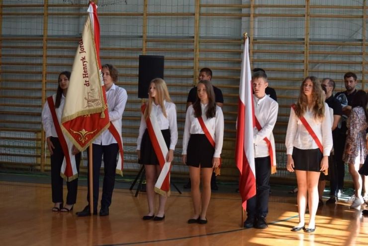Uczniowie ubrani na galowo mają przepasane szarfy w barwach narodowych. Jeden uczeń trzyma sztandar, drugi flagę polski. Obok każdego chłopca stoją dwie dziewczynki