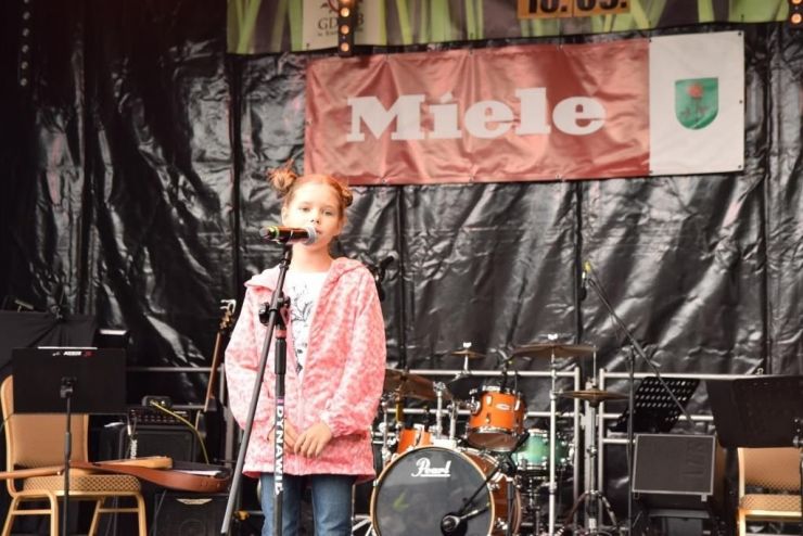 Na scenie stoi dziewczynka w różowej kurtce. Przed nią stoi mikrofon. W tle instrumenty muzyczne