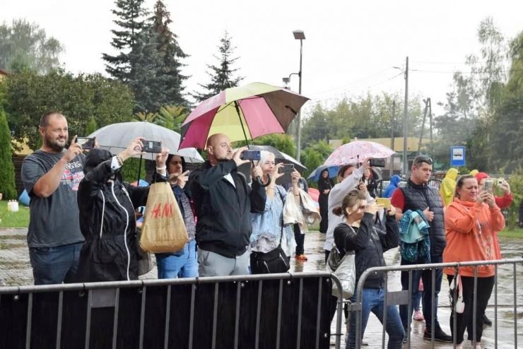Grupa uczestników stoi z parasolami przed sceną. Część osób trzyma telefony komórkowe w ręku i nagrywa występy