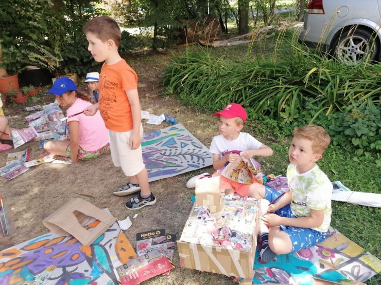 Piątka dzieci jedno stoi reszta siedzi wśród drzew i maluje na dużych fragmentach kartonowych.