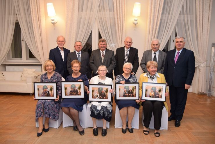  Sześciu mężczyzn stoi przed nimi na krzesłach siedzi pięć kobiet trzymające ramki ze zdjęciami w dłoniach.