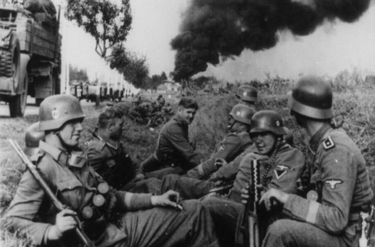 Czarno-białe zdjęcie przedstawiające żołnierzy w hełmach siedzących z bronią na poboczu drogi. W tle widoczny czarny dym