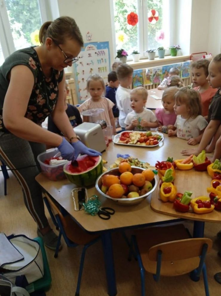 Przy stoliku stoi grupa dzieci i nauczycielka. Na stoliku jest dużo owoców i warzyw. Nauczycielka robi kulki z arbuza