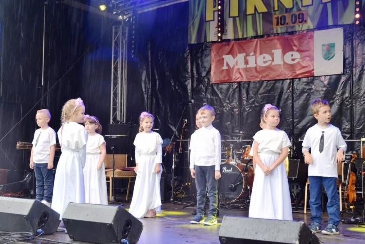 Na scenie stoi grupa dzieci ubranych na biało. W tle widoczne instrumenty muzyczne