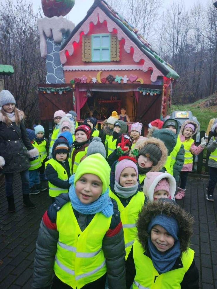 Grupa dzieci w zielonych kamizelkach. W tle kolorowy domek wyglądający jak z bajki