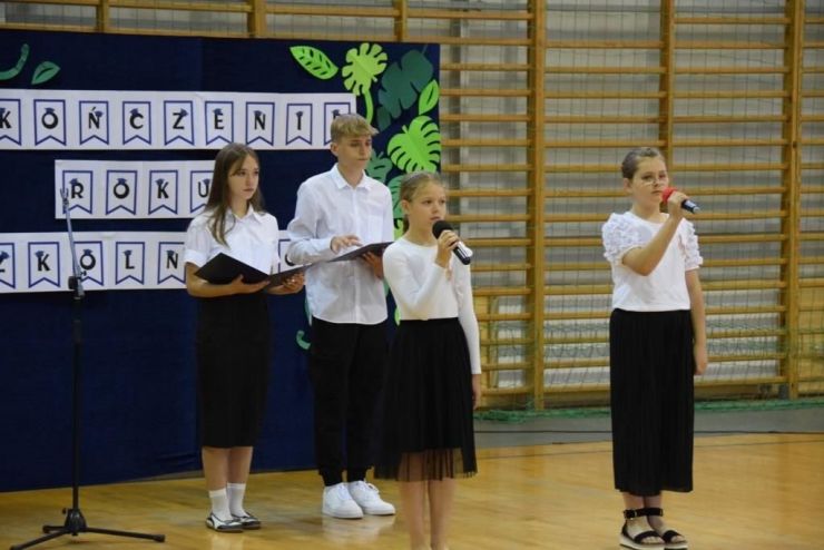 Czwórka dzieci stoi w galowych strojach z mikrofonami przy dekoracji z napisem "Zakończenie roku szkolnego"