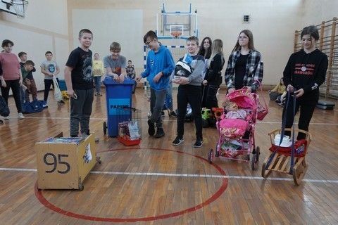 uczniowie stojący na sali gimnastycznej przed nimi wózek drewniany, wózek dla lalki, sanki klatka dla ptaka oraz pojemnik na papier