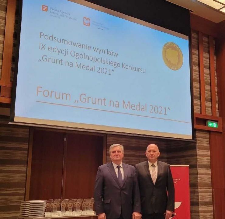 Dwóch mężczyzn w garniturach stoi przy ekranie projekcyjnym na którym widnieje napis "Podsumowanie wyników IX edycji Ogólnopolskiego Konkursu Grunt na Medal 2021